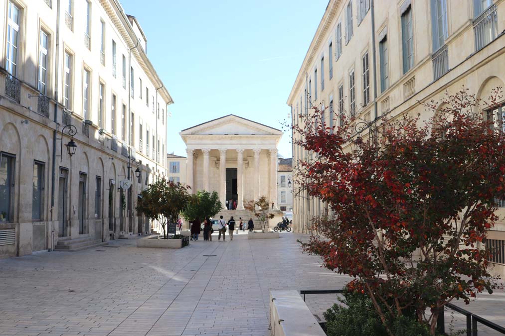 The Parthenon in Nimes, France. Paved pedestrian space with stone building to each side in the foreground.