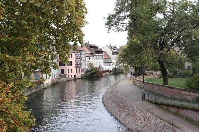Canal in Strasburg, France with path on left side and three storey homes on the right. Trees in foreground.