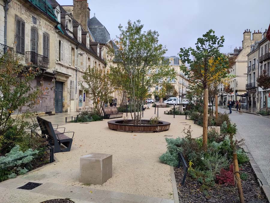 Square in front of three story buildings with gravel, shrubs, trees, benches and bike racks.